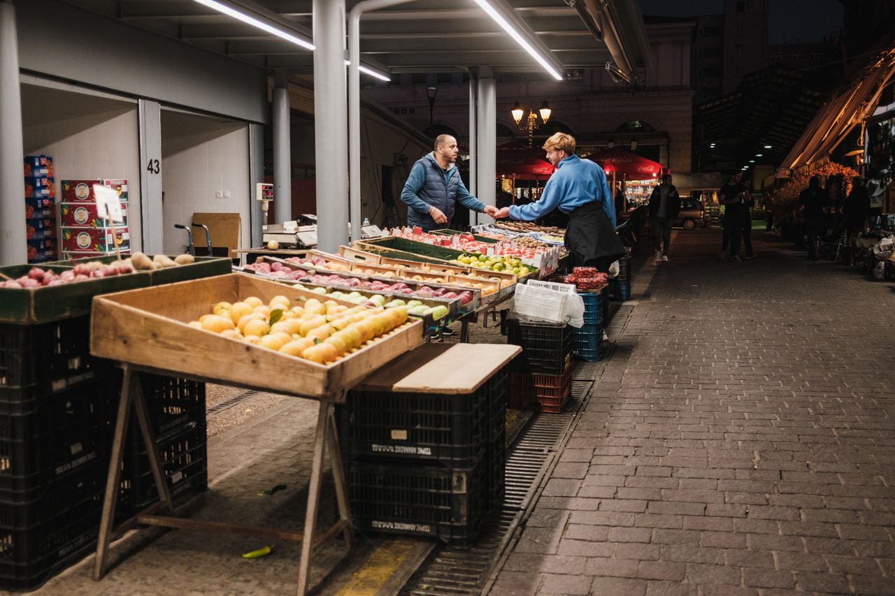 collection-04 Two people exchanging goods at a well-lit night market in the city, surrounded by fresh produce.